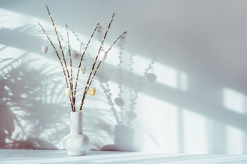 Easter home decor. Modern minimalist style interior with blooming branches of the pussy willow and hanging easter eggs in ceramic vase on a white console with sunlight and shadows on white gray wall.