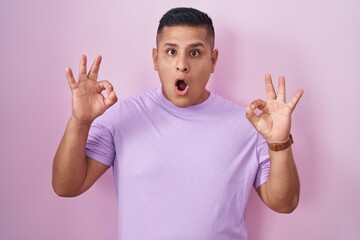 Young hispanic man standing over pink background looking surprised and shocked doing ok approval symbol with fingers. crazy expression