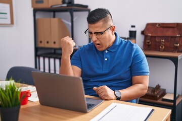 Young hispanic man working at the office with laptop angry and mad raising fist frustrated and furious while shouting with anger. rage and aggressive concept.