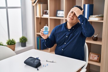 Hispanic young man holding glucometer device stressed and frustrated with hand on head, surprised...