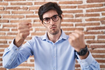 Young hispanic man standing over brick wall background angry and mad raising fists frustrated and furious while shouting with anger. rage and aggressive concept.