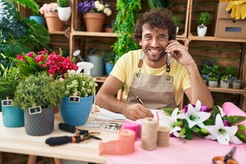 Young hispanic man florist talking on smartphone writing on notebook at flower shop