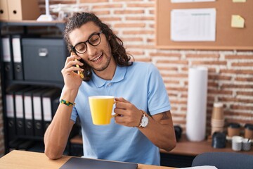 Young hispanic man business worker talking on smartphone drinking coffee at office