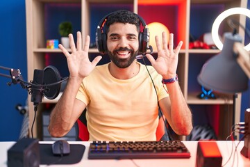 Hispanic man with beard playing video games with headphones showing and pointing up with fingers number ten while smiling confident and happy.