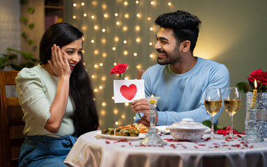 Happy young man proposing excited girl with rose and greetings during candle light dinner - concept...