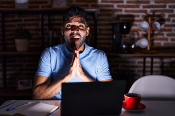 Fototapeta na wymiar Hispanic man with beard using laptop at night begging and praying with hands together with hope expression on face very emotional and worried. begging.