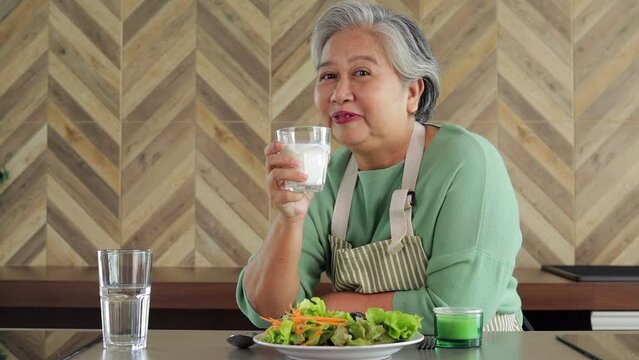 Asian healthy senior woman cooking simple food in kitchen at home in morning time. She drinks skim milk. High calcium helps slow down the occurrence of osteoporosis. Nutritious food for the elderly