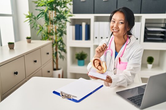 Young chinese woman wearing doctor uniform holding anatomical model of uterus with fetus at clinic