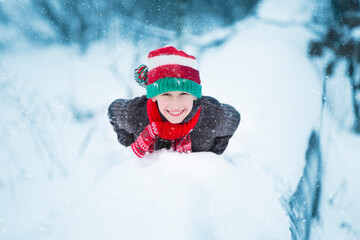 A happy boy in a Christmas hat lies on the snow and smiles.