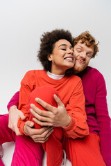 happy redhead man in magenta color sweatshirt hugging cheerful african american woman on grey.