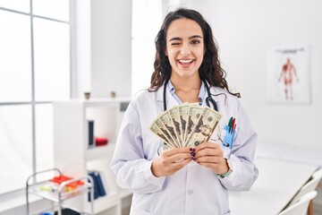 Young doctor woman holding money winking looking at the camera with sexy expression, cheerful and...