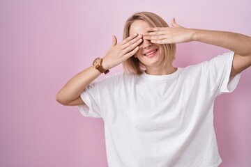 Young caucasian woman standing over pink background covering eyes with hands smiling cheerful and...