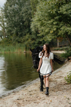 A beautiful girl in a white dress leads a horse along the shore
