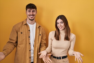 Young hispanic couple standing over yellow background smiling cheerful with open arms as friendly welcome, positive and confident greetings