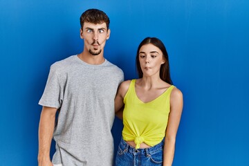 Young hispanic couple standing together over blue background making fish face with lips, crazy and comical gesture. funny expression.