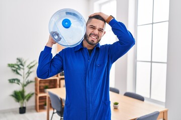 Fototapeta na wymiar Hispanic service man holding a gallon bottle of water for delivery stressed and frustrated with hand on head, surprised and angry face