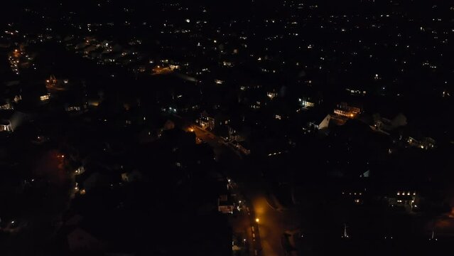Aerial tilt up reveal of houses in large, sprawling American neighborhood during sunset, dusk. Lights on houses, car headlights, and street lamps illuminate dark development. Orange and red sunset.