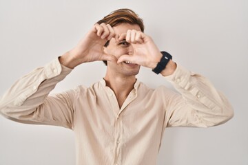 Young man standing over isolated background doing heart shape with hand and fingers smiling looking through sign