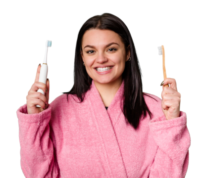 A woman in a pink bathrobe holds both an electric toothbrush and a traditional toothbrush, with a look of uncertainty as she compares the two.