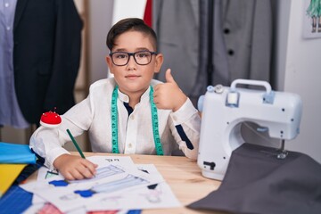 Young hispanic kid at tailor room smiling happy and positive, thumb up doing excellent and approval sign