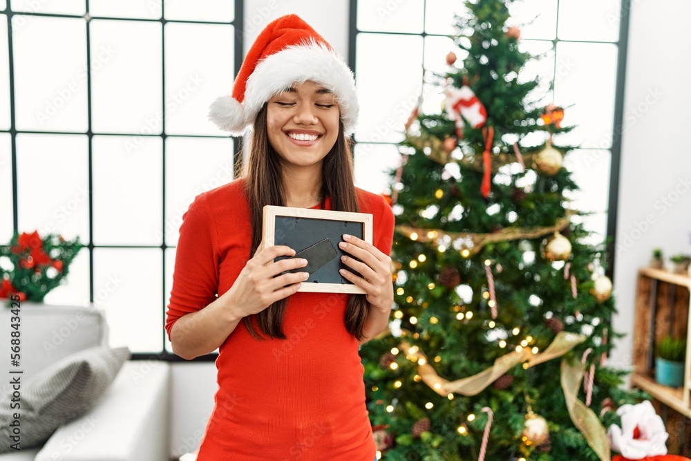 Canvas Prints Young latin woman holding photo standing by christmas tree at home