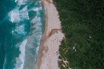 Der Strand Praia da Galheta von oben fotografiert. Viele Sonnenschirme direkt am Meer und daneben Tropischer Wald. Drohne