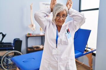 Middle age woman with grey hair working at pain recovery clinic doing bunny ears gesture with hands...