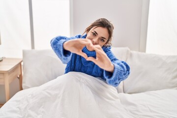 Young beautiful hispanic woman doing heart gesture sitting on bed at bedroom