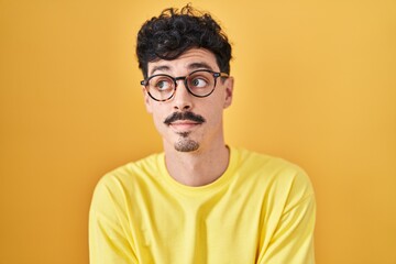 Obraz premium Hispanic man wearing glasses standing over yellow background smiling looking to the side and staring away thinking.