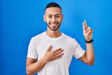 Young hispanic man standing over blue background smiling swearing with hand on chest and fingers up, making a loyalty promise oath
