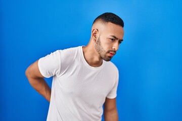 Young hispanic man standing over blue background suffering of backache, touching back with hand, muscular pain