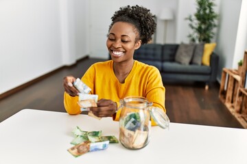African american woman sitting on table holding rand banknotes of charity jar at home