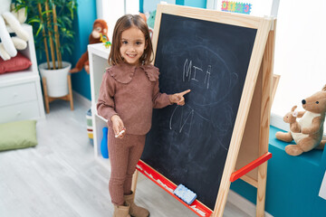 Adorable hispanic girl preschool student smiling confident writing name on blackboard at...