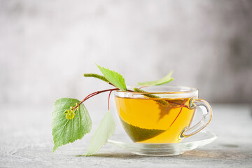 Glass cup of hot, fragrant birch tea on a grey background and a branch of birch leaves
