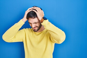 Hispanic man standing over blue background suffering from headache desperate and stressed because pain and migraine. hands on head.