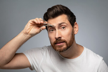 Obraz premium Grooming wellness and facial care. Handsome middle aged bearded man tweezing eyebrows, grey background