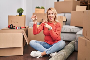 Blonde woman holding keys of new home sitting on the floor smiling happy pointing with hand and finger