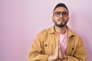 Young hispanic man standing over pink background begging and praying with hands together with hope expression on face very emotional and worried. begging.