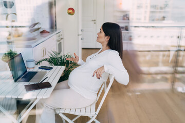 Young pregnant woman tossing apple while sitting at table with computer