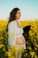 Naklejka na ściany i meble A pregnant woman with a small black dog in nature. Rapeseed field