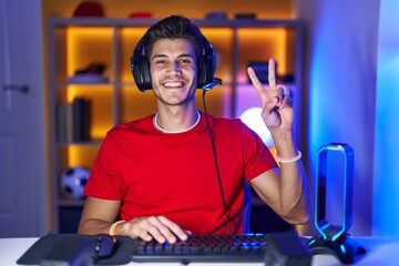 Young hispanic man playing video games showing and pointing up with fingers number two while smiling confident and happy.