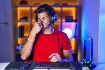 Young hispanic man playing video games smelling something stinky and disgusting, intolerable smell,...