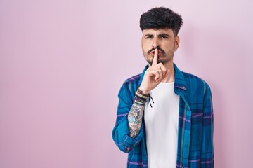 Young hispanic man with beard standing over pink background asking to be quiet with finger on lips....