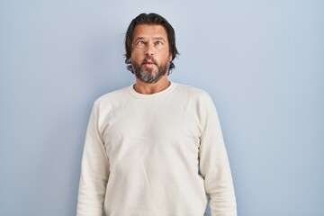 Obraz premium Handsome middle age man wearing casual sweater over blue background making fish face with lips, crazy and comical gesture. funny expression.