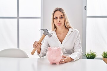 Young blonde woman holding piggy bank and hammer skeptic and nervous, frowning upset because of problem. negative person.