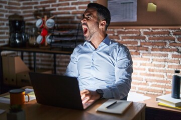 Hispanic man with beard working at the office at night angry and mad screaming frustrated and furious, shouting with anger. rage and aggressive concept.