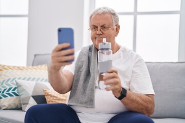 Middle age grey-haired man make selfie by smartphone holding bottle of water at home