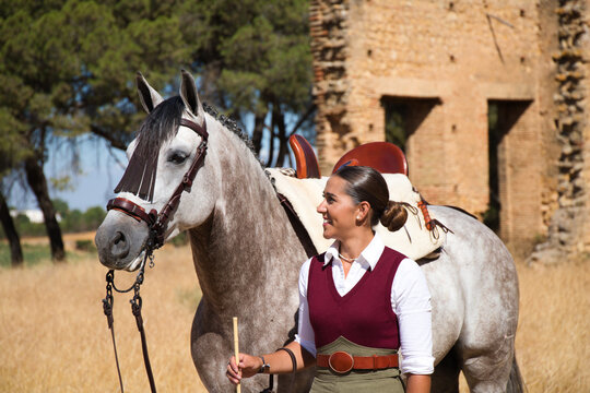 Young and beautiful Spanish woman with her thoroughbred horse. The woman is wearing a typical Spanish riding uniform. They are next to the stable in the countryside in seville. Concept horses.