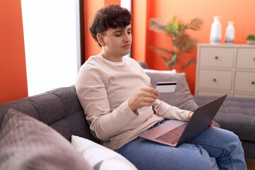 Non binary man using laptop and credit card with worried expression at home