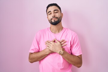 Hispanic young man standing over pink background smiling with hands on chest with closed eyes and grateful gesture on face. health concept.